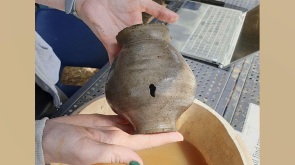 A woman's hands holding up a Roman pot with a hole in its side over a bowl of muddy water