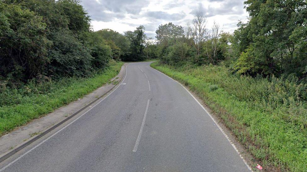 Luton Road - a country B-road with a very thin pavement on the left side and white lines down the middle. Grass, shrubs and trees are on the verges of both sides of the road, which bends round to the right later on.