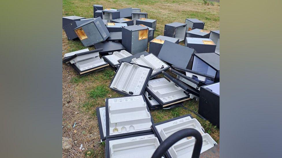 The fridges, dumped on the grassed area. Most of the doors have separated from the bodies of the fridges. They are a dark grey colour.