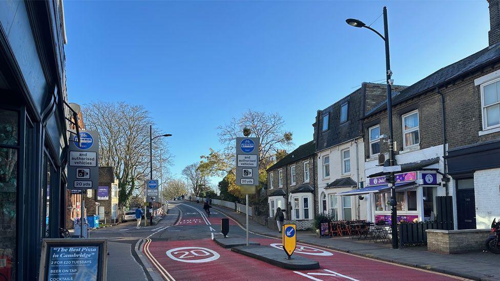 Mill Road bridge in Cambridge. Red road markings show it is a 20mph zone. Shops and cafes are on either side of the road.