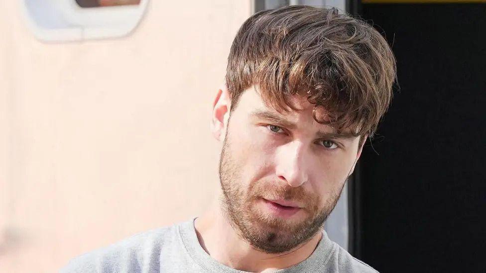 A man with medium length, messy brown hair. He is looking at the camera and wearing a grey jumper