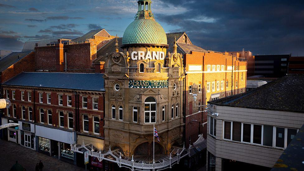 Street view image of the theatre. It is on a corner with red brick buildings to either side and a round stone entrance in the middle with a round, green, copper dome on top