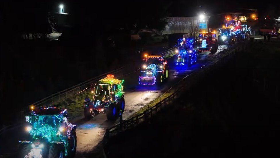 A line of illuminated tractors drive up a dark road. They are illuminated with loads of different coloured lights.