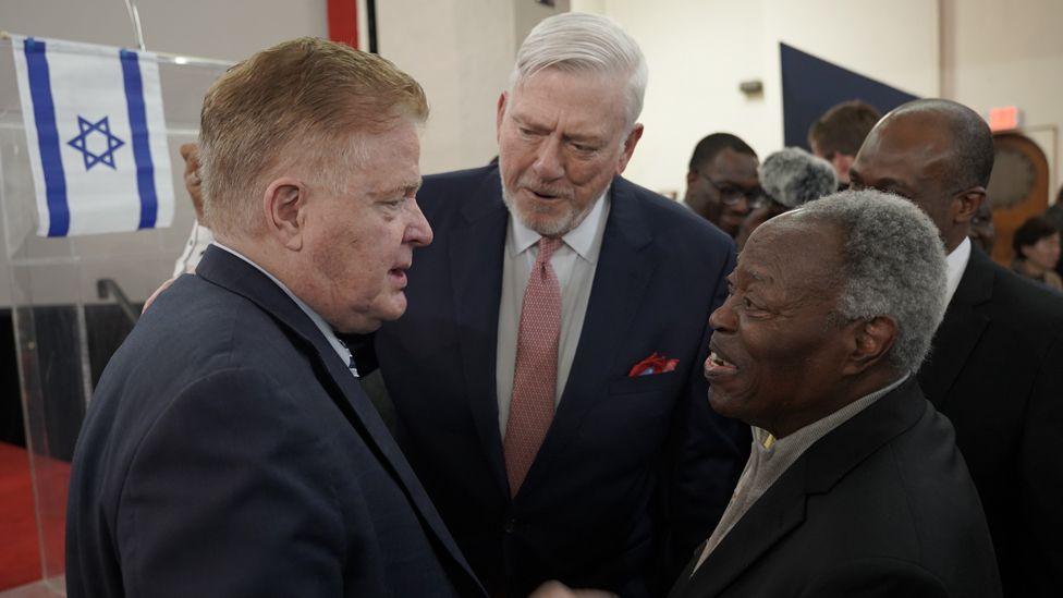 William Kumuyi in conversation and shaking hands with Jim Garlow next to a lectern that has the Israeli flag on it.