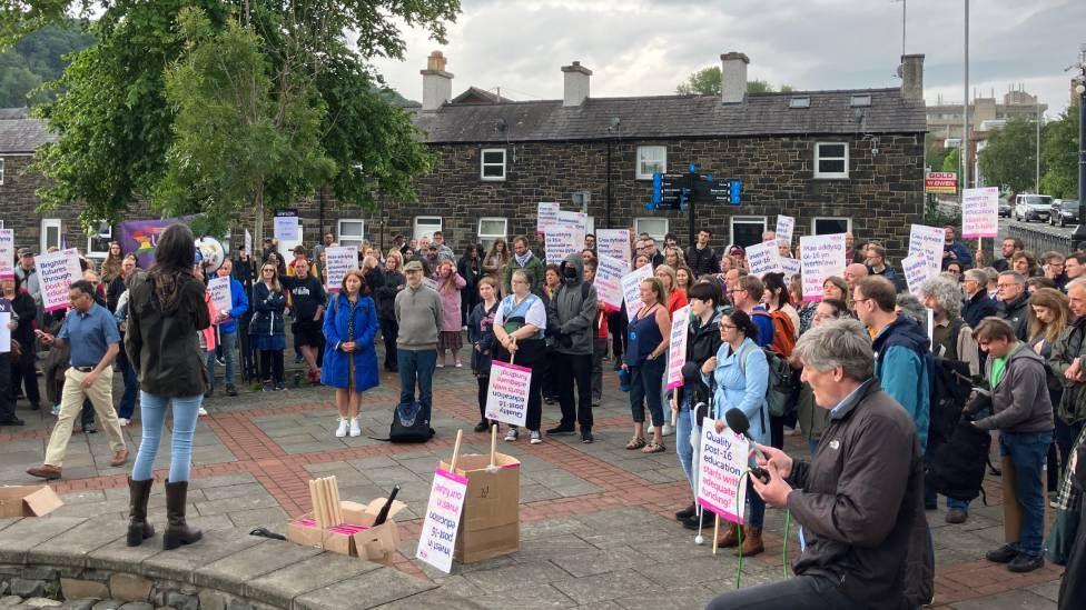 A crowd of people holding pink and white placards with writing on them. Behind them are some houses and to the left is a tree. Standing to the left, on a bench, addressing the crowd is a woman with dark hair, wearing a black jacket and light blue jeans