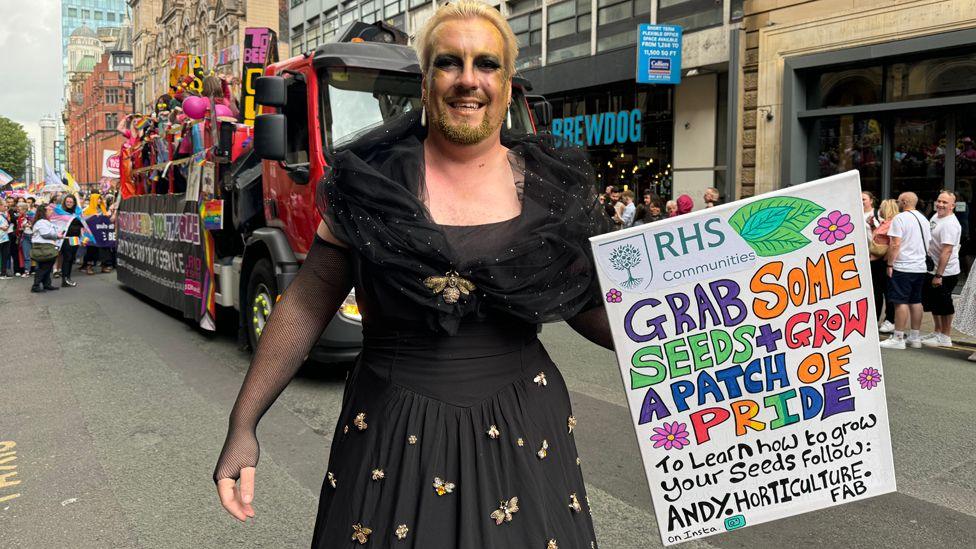 A man with a gold-painted face and beard and wearing a black dress with gold bee decorations carrying a sign which has a message about "growing a patch of Pride" from seeds in the Manchester Pride parade