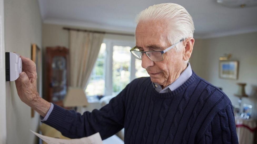 An older man adjusting a thermostat on a wall. He is wearing a blue jumper over a shirt.