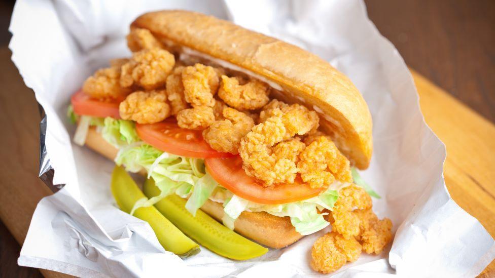A po' boy sandwich, made with fried shrimp, tomato, lettuce, pickles and sauce, is served on a wrapper on a wooden table