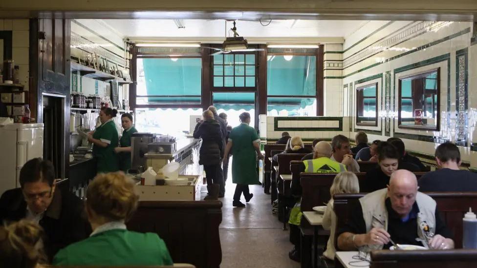 People sitting at tables and eating in a cafe. Waitress wearing green dresses are standing and walking around.