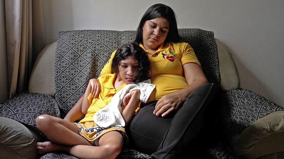 Rute (right) wears a yellow polo shirt while sitting on a sofa. She has her arm around her daughter Tamara, who holds a white towel and has dark curly hair.