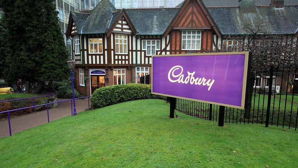 The front of the Cadbury headquarters in Bournville. A large purple sign with the word Cadbury sits at the front of the grass lawn, with a large building behind.