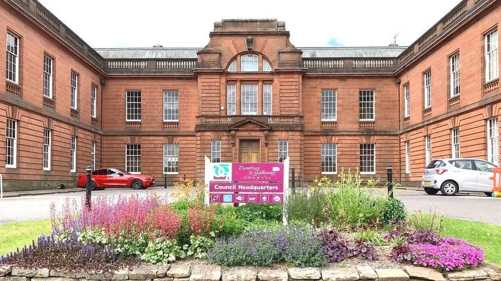 Dumfries and Galloway Council headquarters is a large sandstone building with rows of long white framed windows. The front of the building has threes sides with a car park and floral display in the middle.