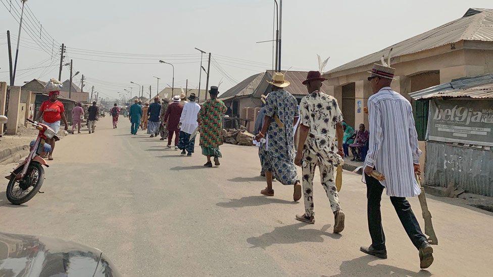 Members of the Igbuu Society seen walking down a street in Oguta in single file each holding a gold sword