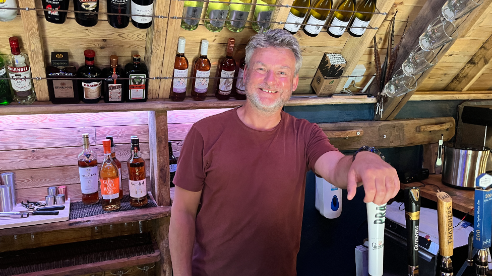A man is stood with his hand on a beer tap while stood behind a bar looking at the camera.