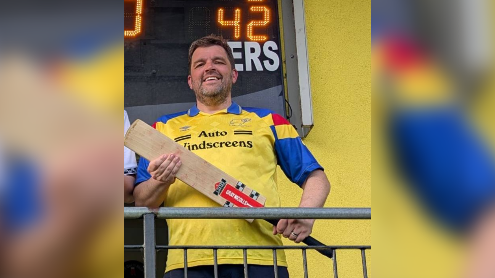 Ian Bestwick in a yellow cricket shirt. He holds a bat and stands in front of a scoreboard.