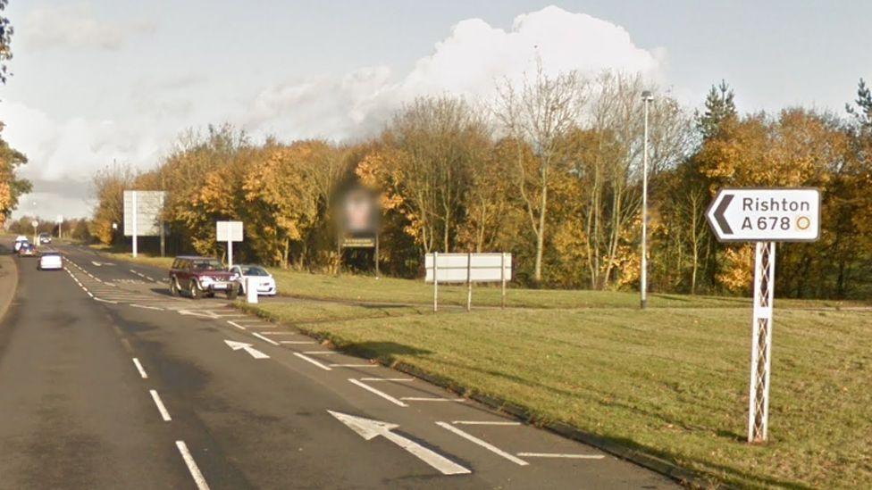 Generic street view of the end of Blackburn Road in Rishton, showing a sign pointing to the left saying "Rishton A678", on a grassed area. There are trees in the background as two cars drive towards a roundabout