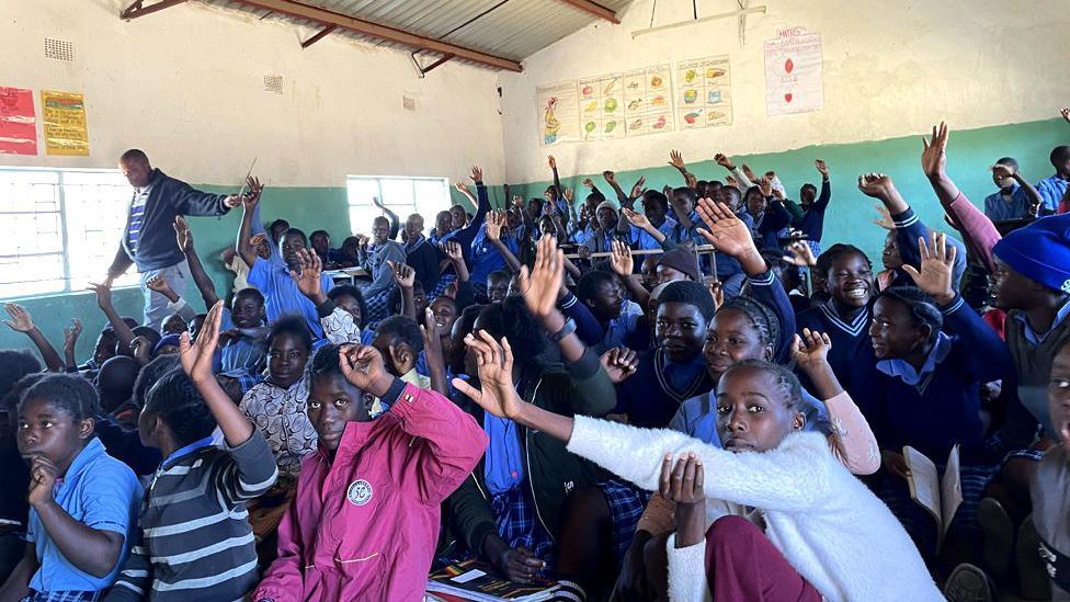 A packed classroom with pupils holding their hands up 