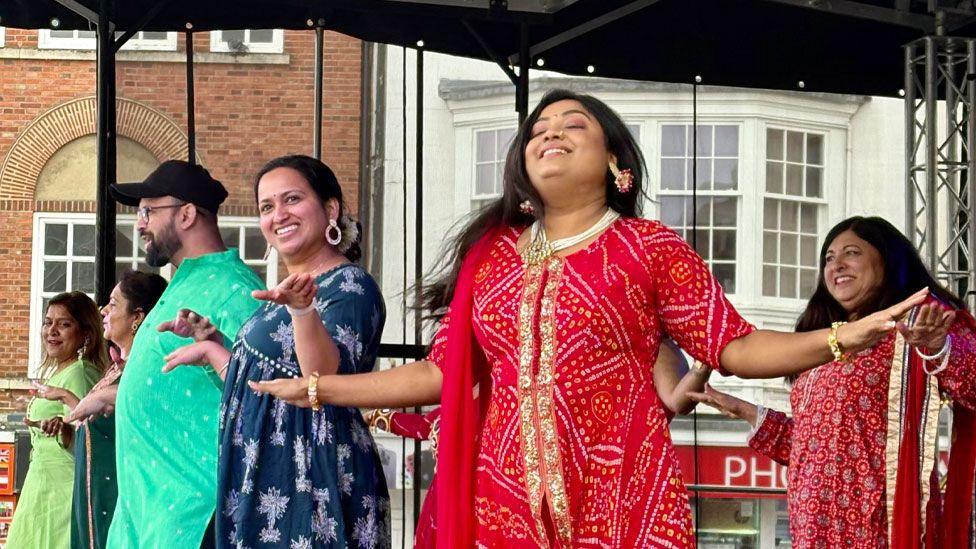Five women and one man dancing on a stage in Northampton's Market Square. They are wearing brightly coloured Indian clothing and are smiling and laughing.