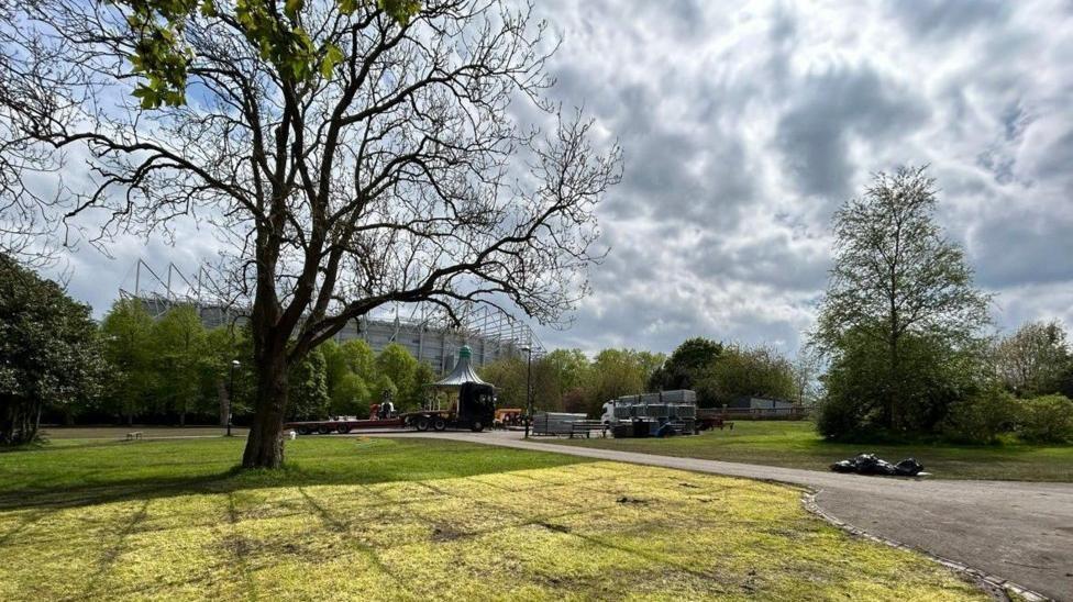 A view of Leazes Park, with St James' Park in the background. In the foreground there is a patch of grass with a leafless large tree. There is a path to the right that leads to a bandstand which has a dark lorry parked in front of it.