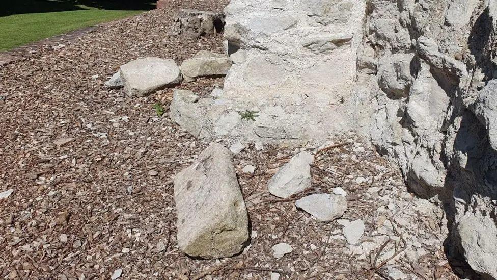 A close-up of the foot of a ruined ancient castle wall. Several large pieces of stone are pictured lying next to an intact wall, indicating these have been moved or pushed off the top of the wall by vandals.