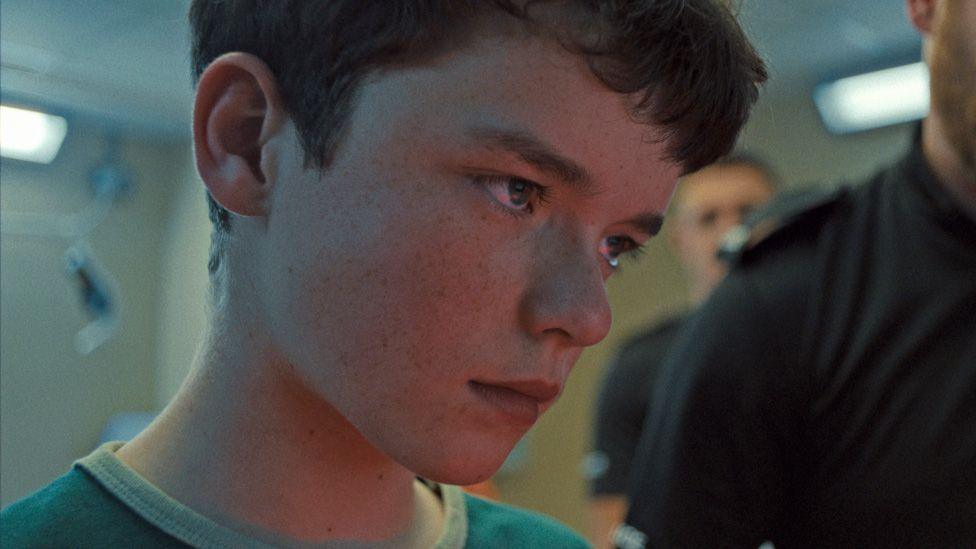 Owen Cooper, who plays Jamie, stands in a police station. He is staring off to the side. He wears a blue T-shirt and has short dark hair.