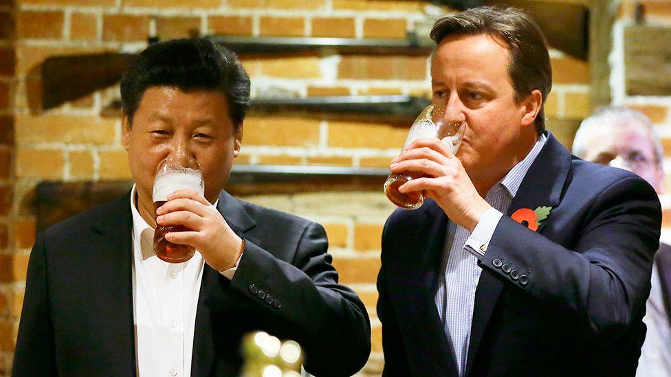 Xi Jinping and David Cameron wearing suits and open-necked shirts, holding and drinking pints of beer in a rustic setting with exposed brick walls and mounted antique rifles in the background.