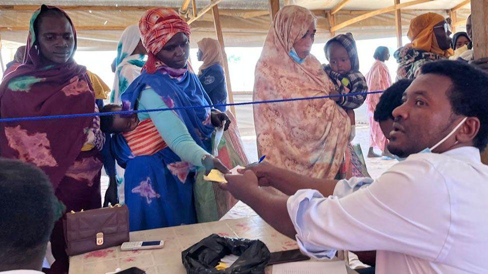 A seated aid worker in Chad looks after his shoulder as he hands paperwork to a woman who is in a line of newly arrived people from Sudan