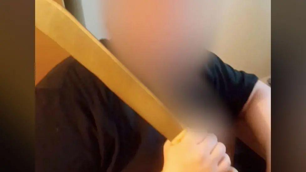 A heavily blurred photo of X, who is wearing a black T-shirt and holding a large machete