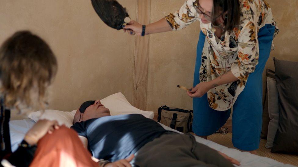 A blindfolded man in a blue T-shirt lies on a bed as a woman stands over him fanning feathers from the wing of a bird.