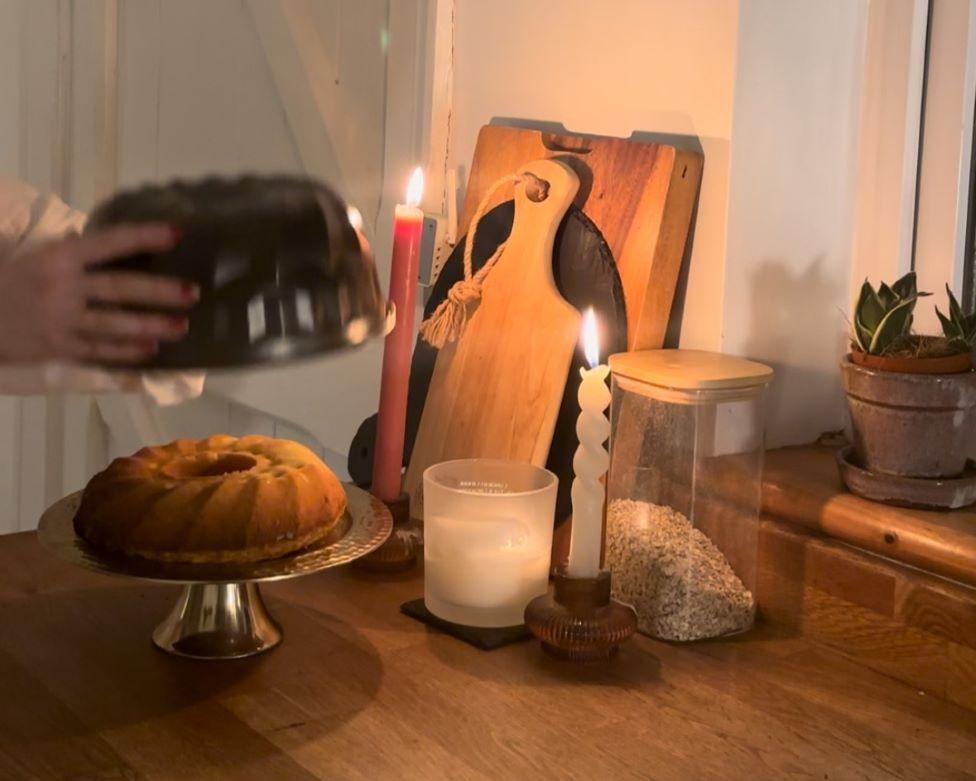 A wooden kitchen counter top containing a Bundt cake, three candles, a jar of oats, three chopping boards and a plant in a pot. A pair of hands are removing the Bundt cake from the tin