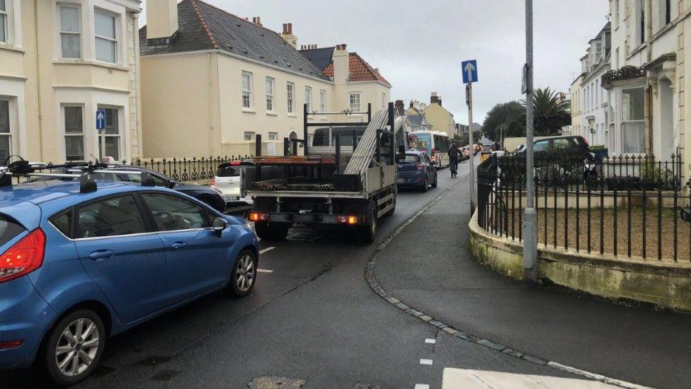 A number of vehicles in a narrow town street, including cars and a small truck carrying scaffolding