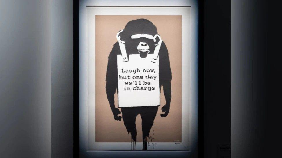 A Banksy artwork of a money with a white sign around its neck reading "laugh now, but one day we'll be in charge"