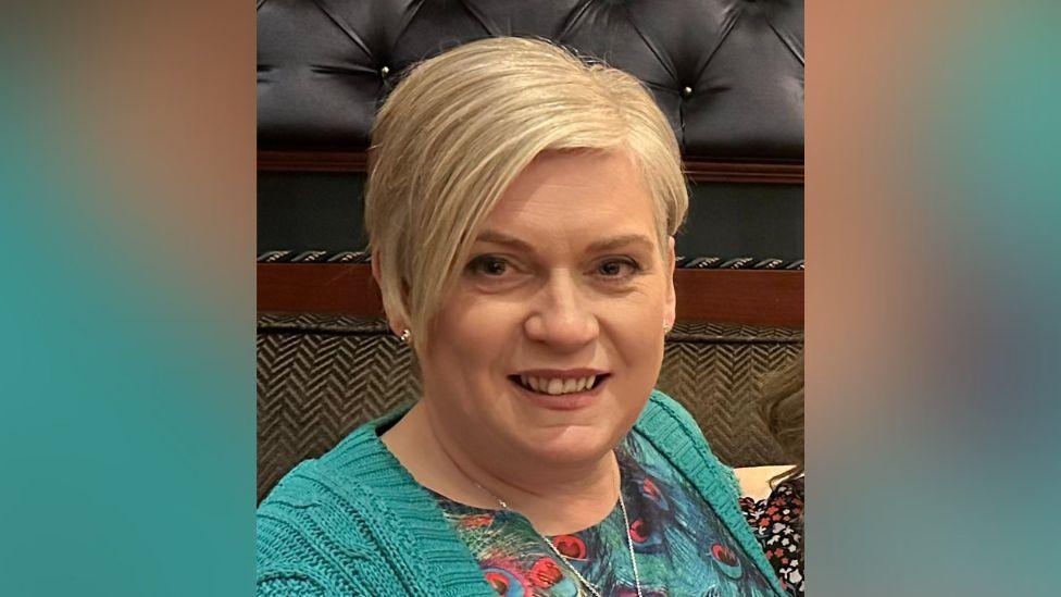 A lady with short blond hair smiles. She is wearing a green cardigan and green and red blouse. She has silver earrings an a silver necklace