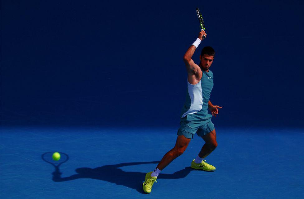 Tennis player Carlos Alcaraz hits the ball during his fourth-round match against Britain's Jack Draper at the 2025 Australian Open. He is wearing a teal outfit and neon yellow shoes on a bright blue court, with his shadow appearing as though it is hitting the ball.