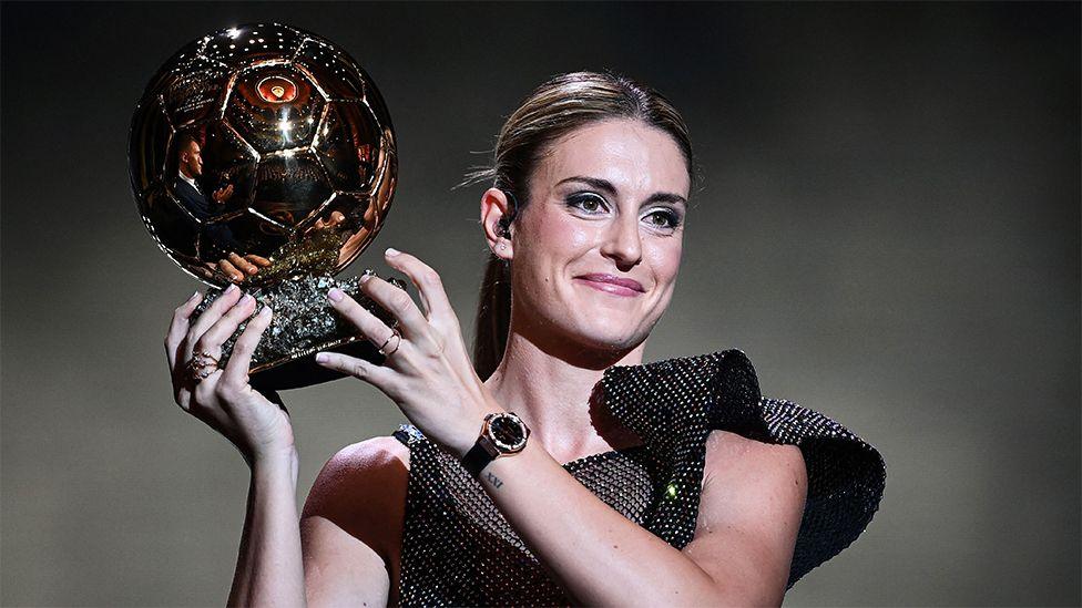 Alexia Putellas holding aloft a football shaped trophy, while dressed in a black glittering outfit.