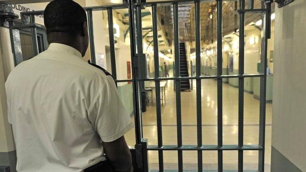 A prison officer in his white prison shirt standing with his back to the camera looking through a prison barred door at the entrance of a prison corridor 