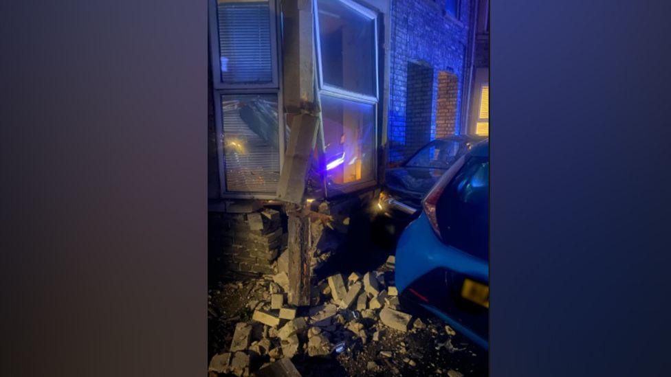 Damage to the front bay window of a brick house shows masonry falling off and a pile of bricks on the pavement. Two cars can also be seen.