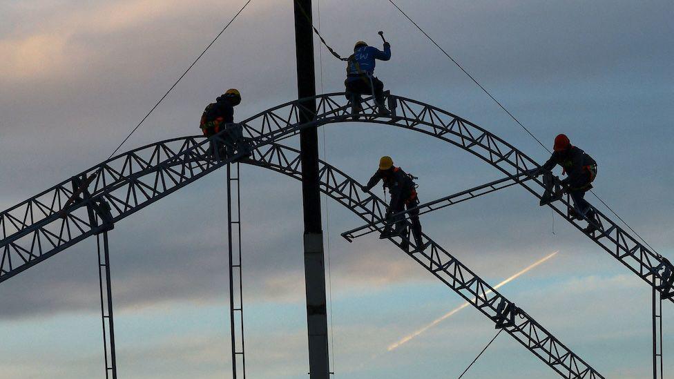 Construction workers are silhouetted against a pale blue sky as they start to set up a giant tent in Ciudad Juarez