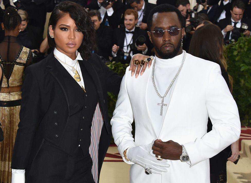 Cassie Ventura and Sean Combs posing for photographers at the 2018 Met Gala. She's wearing a black suit and he's wering an all-white suit with one white glove and a large jewelled cross on a chain around his neck