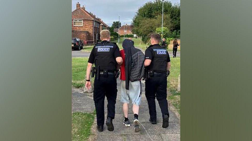 Two police officers dressed in black holding a man by his arms and leading him down a residential street. He's dressed in shorts and a red t-shirt and covering his head with a tracksuit top.