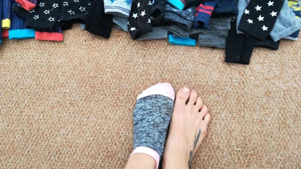 a person's feet but wearing one sock, near a pile of colourful socks