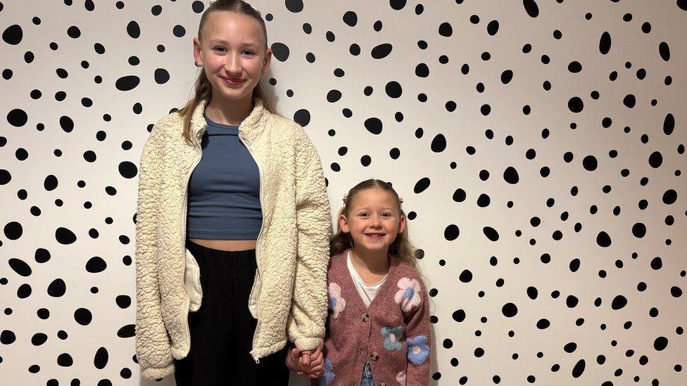 Two young girls stand side by side holding hands - they both have long fair hair - behind them is a black and white spotted wall.
