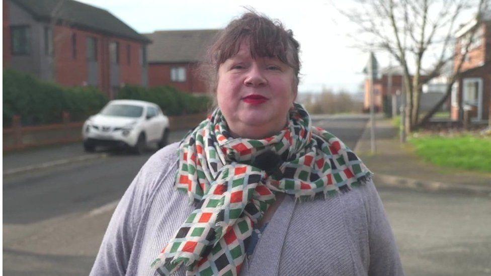 Lucy Antal, with brown hair cut into a fringe and wearing a scarf with a colourful diamond pattern over a grey cardigan, stands on a street looking into the camera 