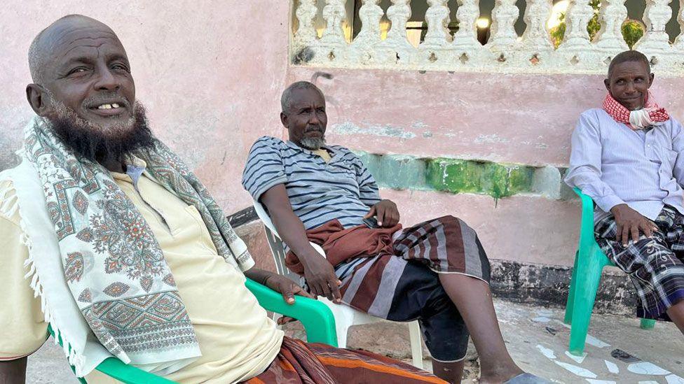 Three men - two with beards - sit on plastic chairs outside in Eyl, Somalia