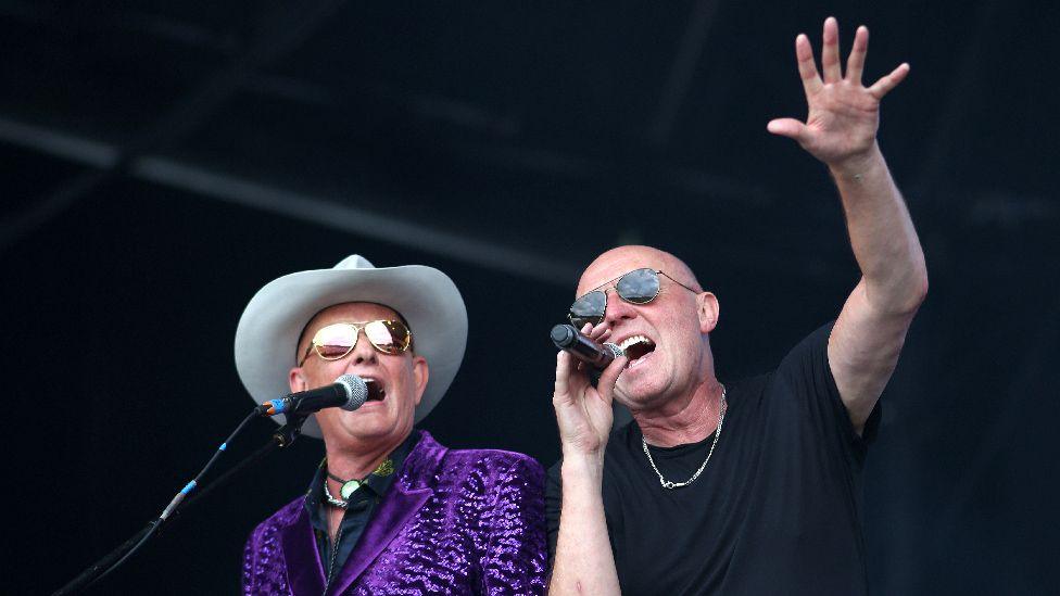 Two men on a stage sing into microphones. One wears a black t-shirt and sunglasses and holds his hand up in the air, fingers spread. His band-mate also wears sunglasses, alongside a cowboy hat and purple, sparkly jacket.