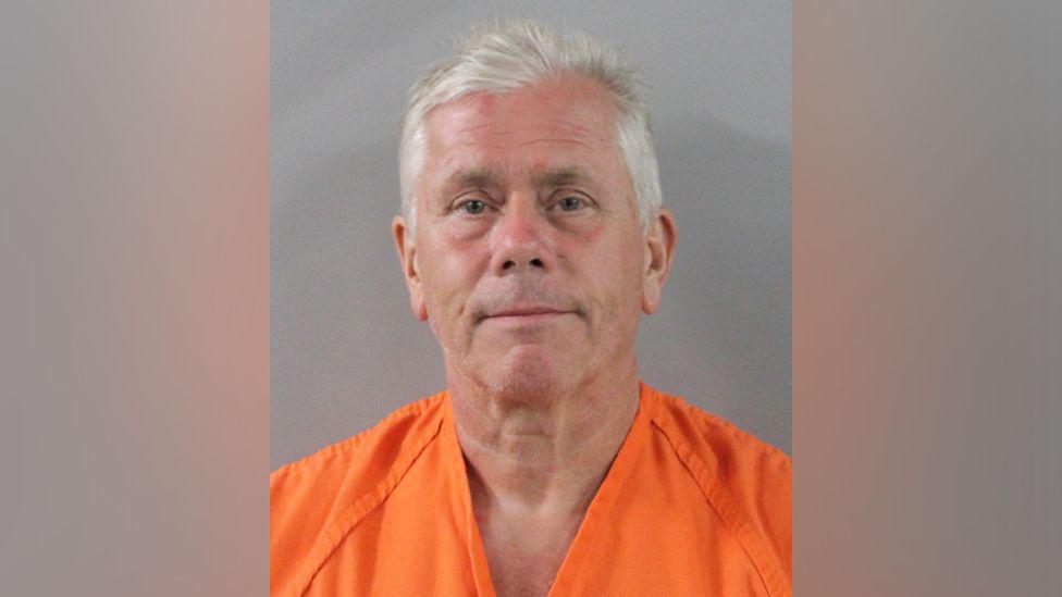 A man in his sixties with white hair looks at the camera during his police mug shot.