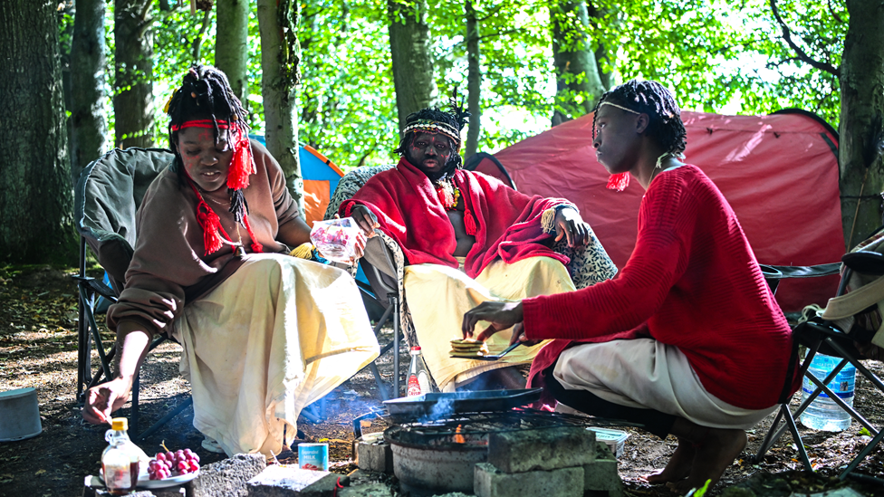 Three people sitting on camp chairs in woodland. A large man in red and cream robes sits at the back. Two women are tending to a fire. There is a red tent in the background.