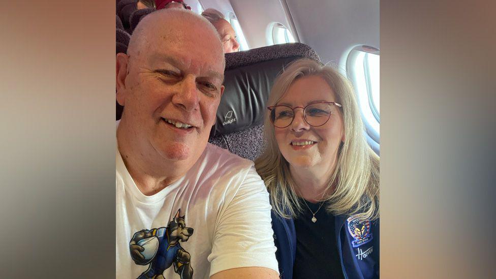 Ged is wearing a white t-shirt with Warrington Wolves wolf illustration on it. Jean is wearing a blue t-shirt and a blue Warrington Wolves jacket and a gold pendant necklace.