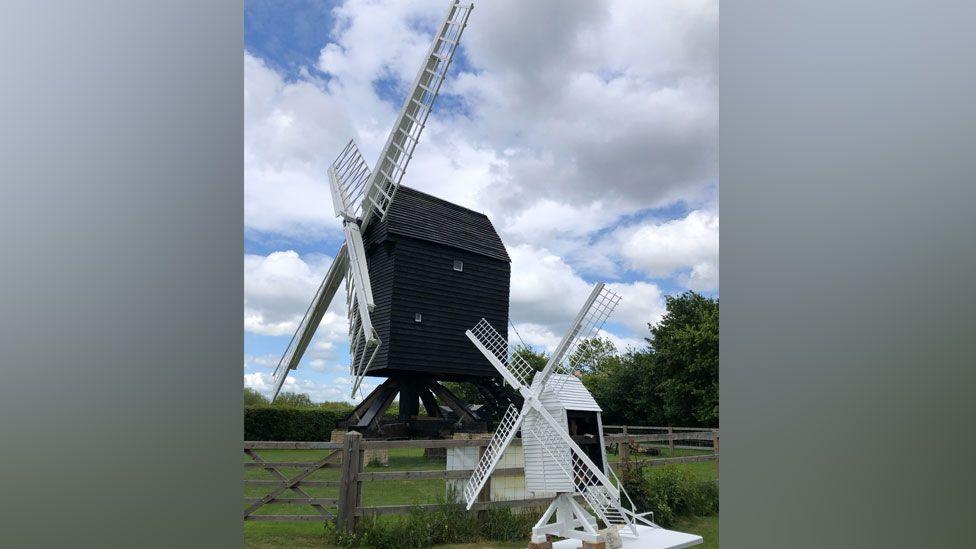 Two wooden mills. The one in the background if a full size black-bodied wooden mill, sideways on with its white sails open in an X shape. The one in front is a white miniature replica, resting on a table. The mills are in fields under white clouds and blue sky. 