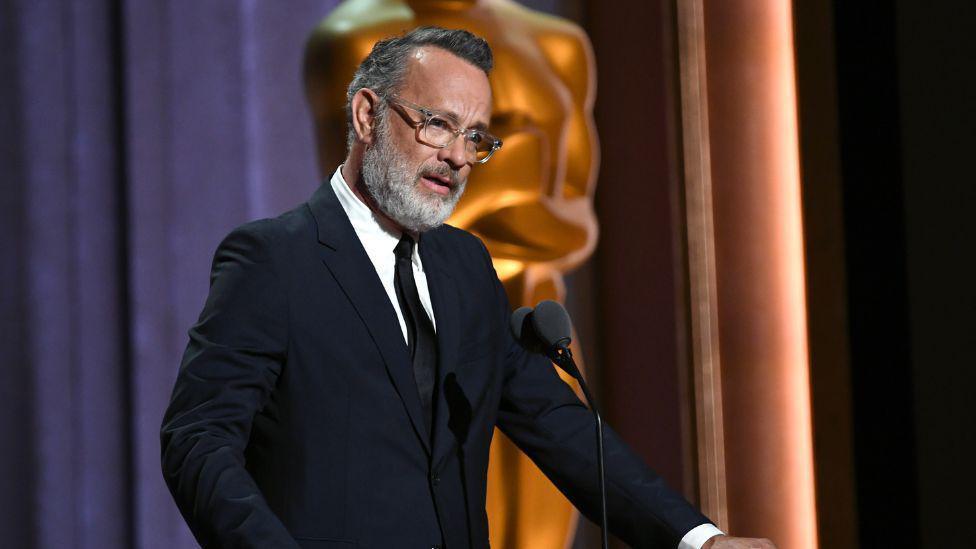 Tom Hanks wearing a black suit and tie, with a white shirt. He is wearing glasses and has grey hair and a grey beard. 